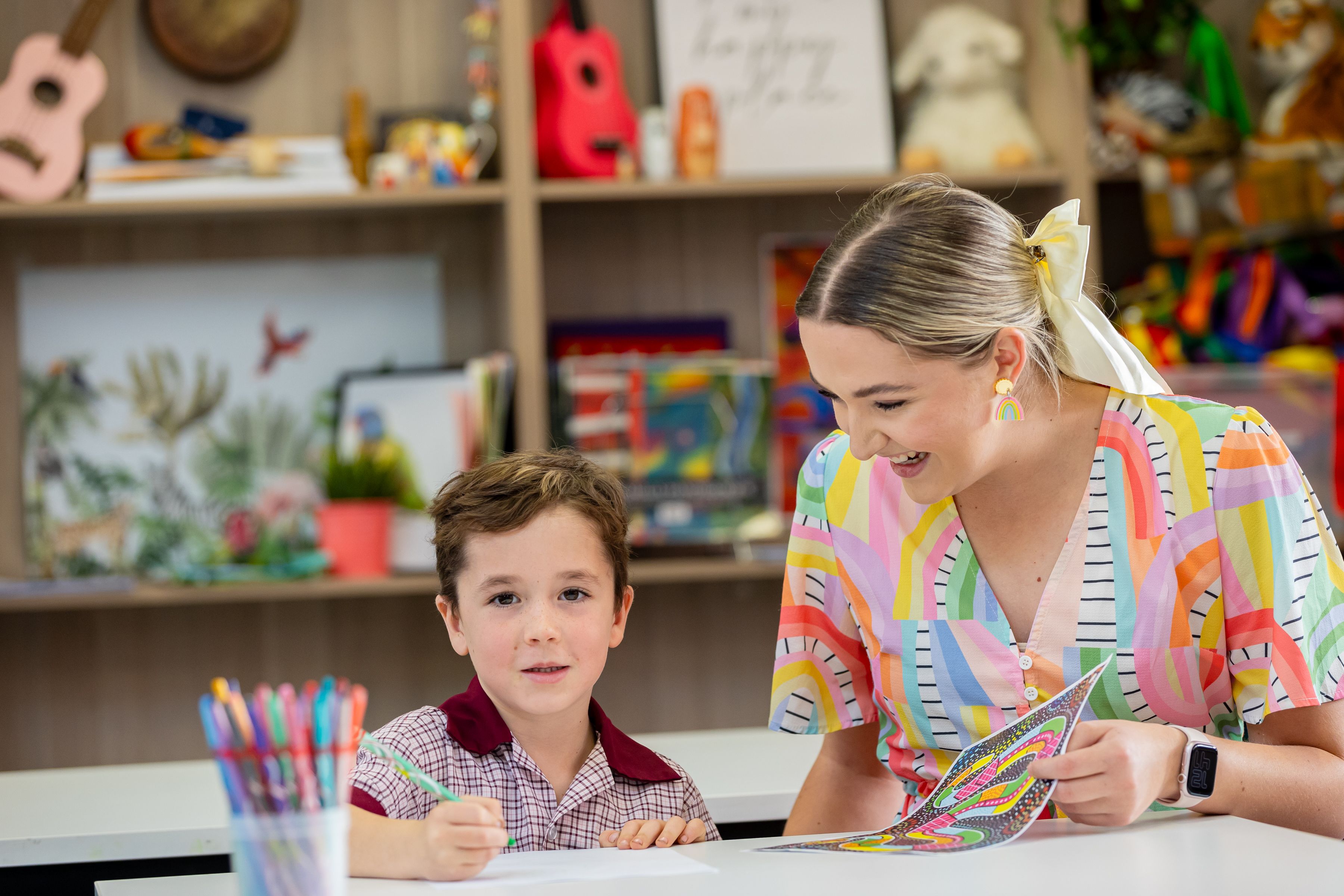 Teacher supporting a student drawing at a desk in a classroom.